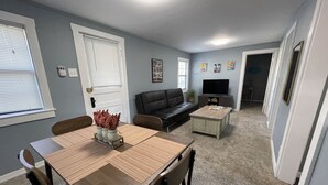 A dining area with a wooden table and chairs, a sofa, and a coffee table. There is a door with a window, a television on a stand, and artwork on the walls. - The Historic McGregor Cottage (Fort Myers)