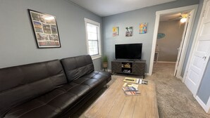 A living room with a brown leather couch, a wooden coffee table with magazines, a TV on a wooden stand, and artwork on the walls. - The Historic McGregor Cottage (Fort Myers)
