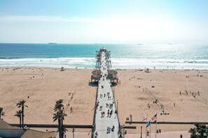 A beach with a long pier extending into the ocean, palm trees, and flags.