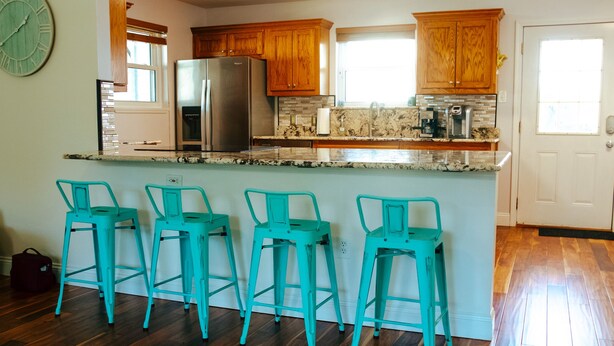 A kitchen with wooden cabinets, a stainless steel refrigerator, a marble countertop, and four turquoise bar stools. - Spacious and stylish 3bd home minutes from beach (Pass Christian)