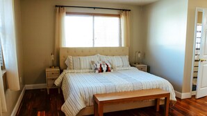 A bedroom with a large bed, wooden floor, bedside tables, and a window with curtains. - Spacious and stylish 3bd home minutes from beach (Pass Christian)