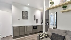 A modern living room with a grey sofa, a coffee table, and a kitchen area with grey cabinets and a stainless steel refrigerator. - Appartement Ricca - Welkeys (Paris)