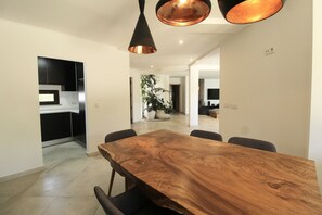 A spacious dining area with a large wooden table and black chairs, a kitchen with black cabinets, and a potted plant in the background.