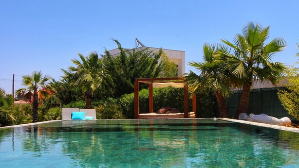 Ein klares Schwimmbecken mit einer Holzpergola und Liegestühlen, umgeben von Palmen und Grünpflanzen.