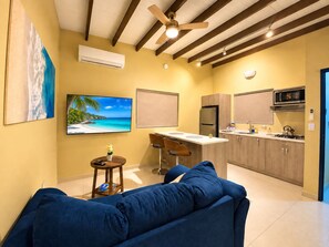 A modern living room with a blue sofa, a TV, and a kitchen area with wooden cabinets and a microwave. - Aruba IXORA A1 - Blenchi Suite, 1-bdrm, sleeps 2, 7 minutes drive to the beach (Noord)
