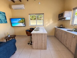 A compact kitchen with a central island, a dining area with chairs, a television mounted on the wall, and a window with a view of palm trees.