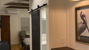 A compact room with a wooden floor, a bed, a desk, and a chair. There is a sliding door leading to another room.