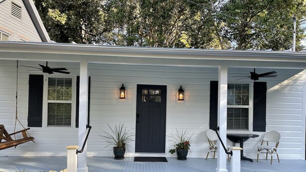 A white house with a black front door, two potted plants, a bench, and a table with chairs on the porch.
