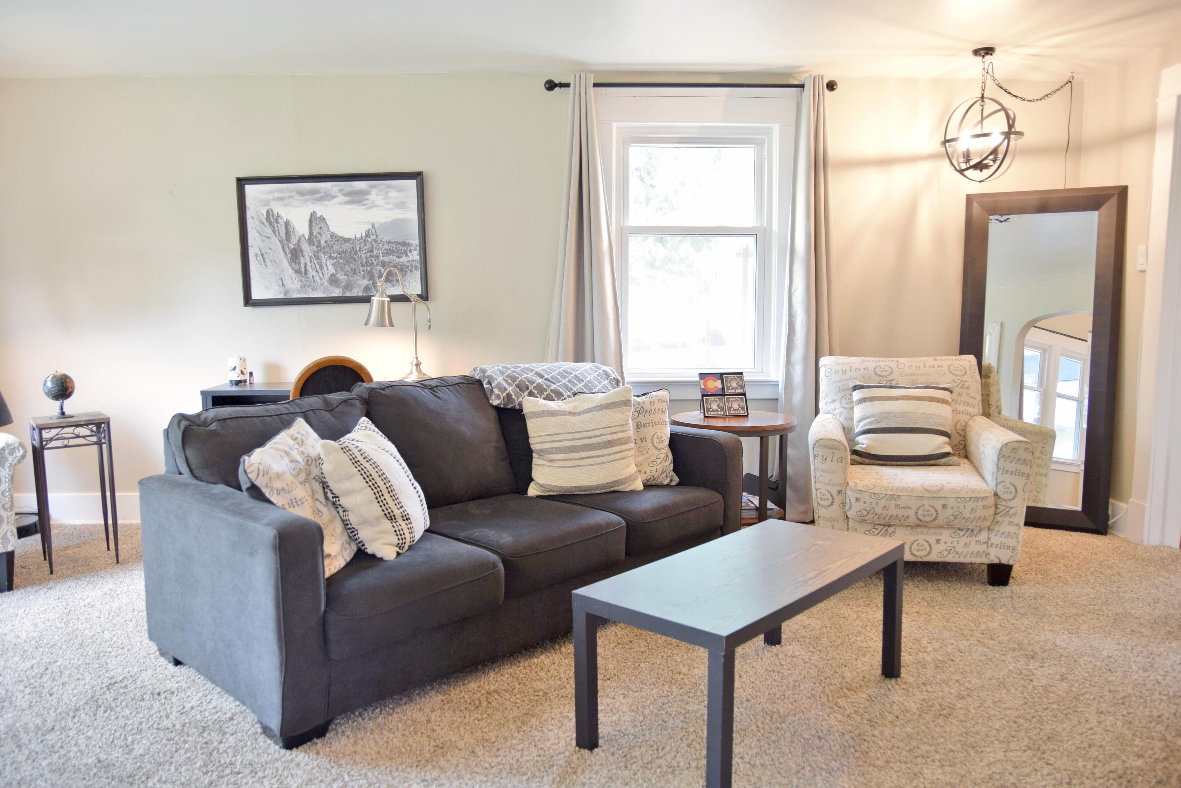 A living room with a dark grey sofa, a small table, a patterned armchair, a framed picture on the wall, and a window with curtains.