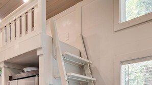 Una cocina con un refrigerador de acero inoxidable, una escalera blanca que conduce a un espacio de almacenamiento elevado, y una ventana con persianas.