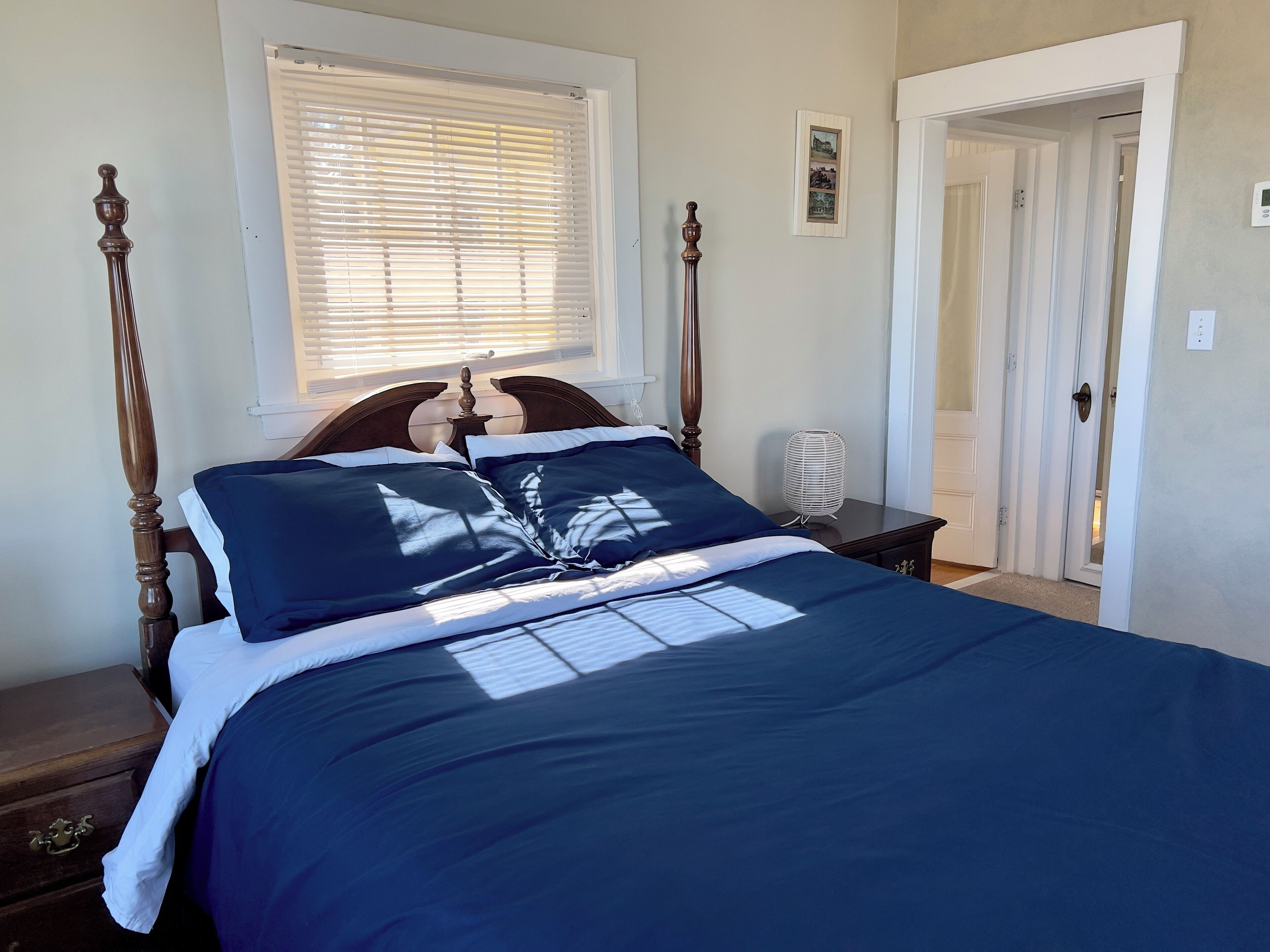 Una cama bien hecha con edredón azul oscuro y almohadas blancas, cabecero de madera, mesita de noche con lámpara y ventana con persianas.