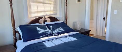 Una cama bien hecha con edredón azul oscuro y almohadas blancas, cabecero de madera, mesita de noche con lámpara y ventana con persianas.