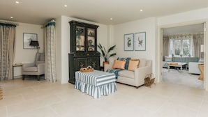 Amplia sala de estar con un mueble de madera oscura grande, un reposapiés a rayas, un sofá color beige con cojines de colores y vista a un área de comedor contigua.