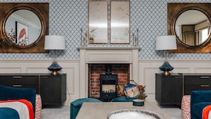 Un salón con chimenea, dos armarios de madera oscura, un sofá azul, un reposapiés con estampado rojo y blanco, y un cuenco grande sobre una mesa.