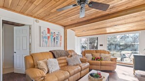 A spacious living room with a large brown leather sofa, a smaller sofa, and a coffee table. There is a ceiling fan, a painting on the wall, and a large window providing a view of the outdoors.