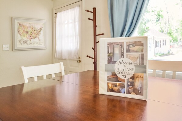 Une table à manger en bois, ornée d’un panneau d’accueil encadré, une chaise blanche, une carte accrochée au mur et une fenêtre avec des rideaux.