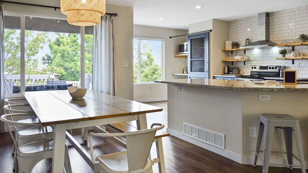 A modern kitchen with a wooden dining table, white chairs, a hanging light fixture, and a view of greenery outside. - Spacious 4 Bed, Central AC, Fire Pit, Grill, Games (Colorado Springs)