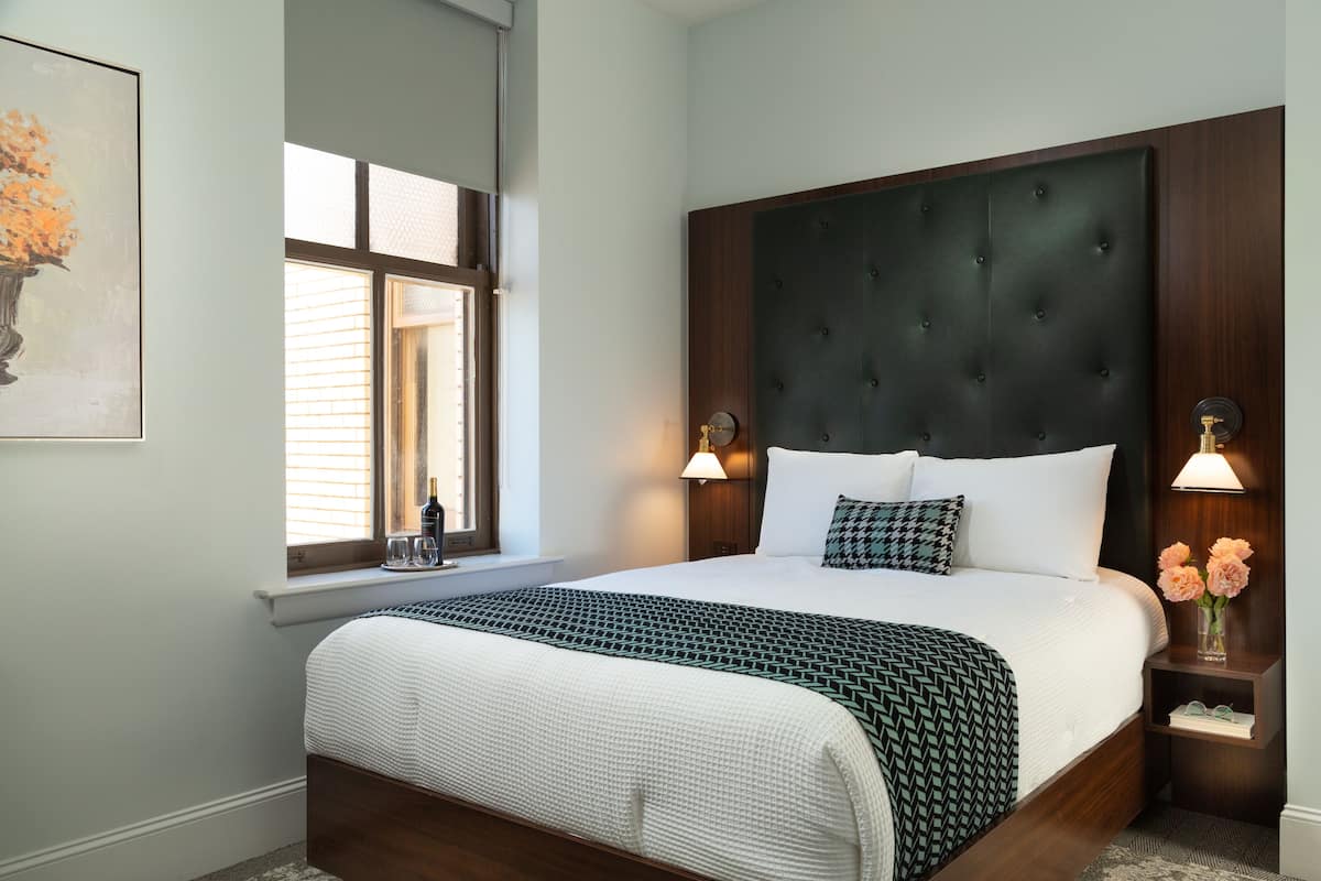 Una cama bien hecha con un cabecero oscuro, dos lámparas de noche y un florero con flores en una mesita de noche.