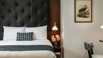 Una cama bien hecha con ropa de cama blanca y estampada, un cabecero capitonado oscuro, una mesita de noche con una lámpara, una ilustración enmarcada de vida silvestre y una silla.