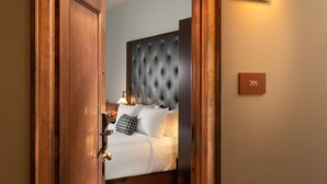 A hotel room with a wooden door, a bed with white bedding, a tufted headboard, and a wall-mounted light fixture. - The Liberty Trust (Roanoke)