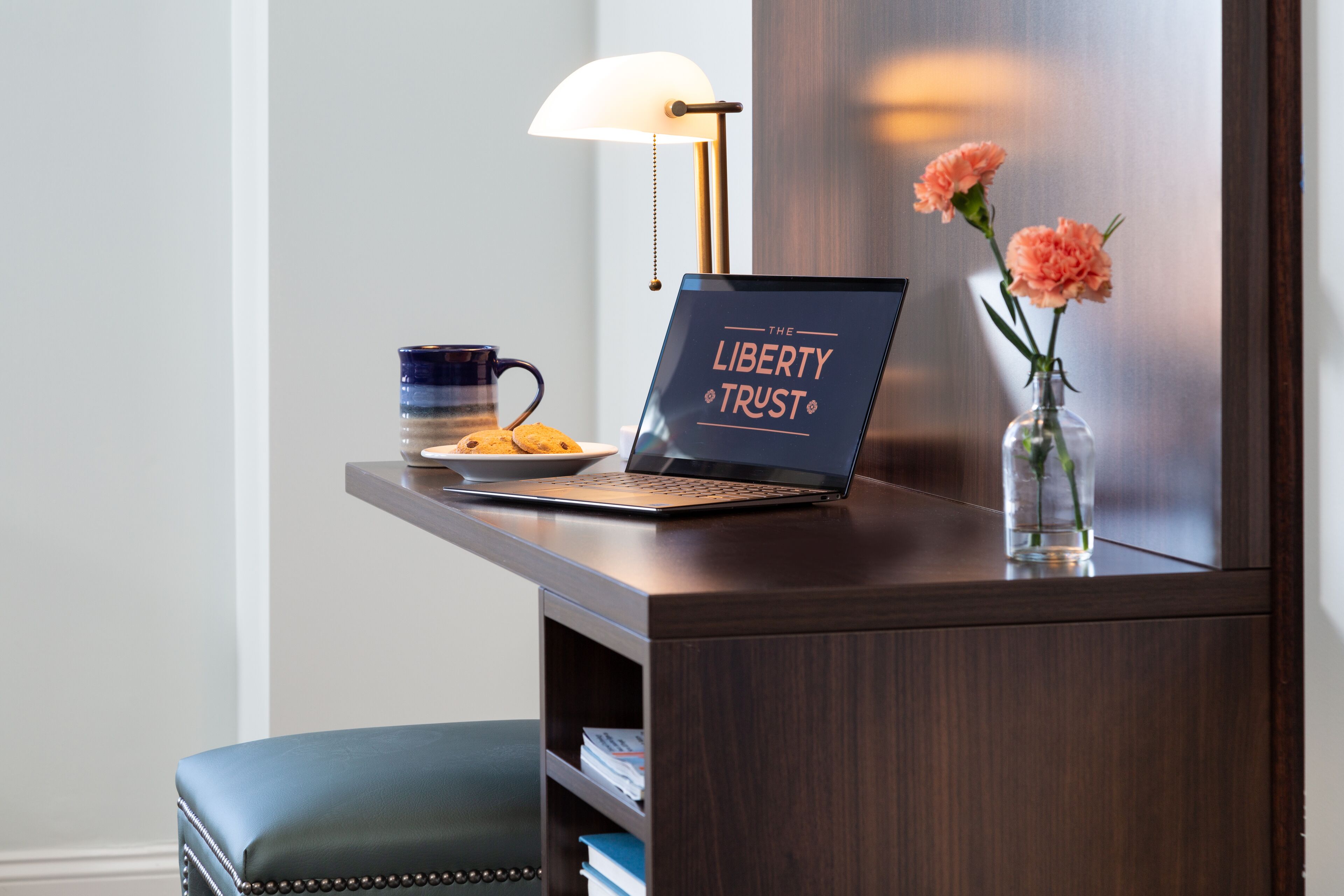 Un ordinateur portable posé sur un bureau, avec une pancarte indiquant « The Liberty Trust », une tasse, une assiette de biscuits et un vase rempli de fleurs.