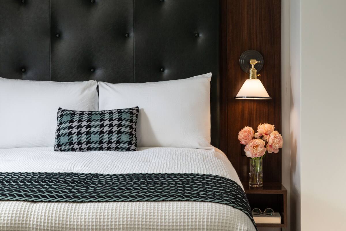 Una cama bien hecha con una colcha estampada, un cojín de pata de gallo y una mesita de noche con un jarrón de flores.