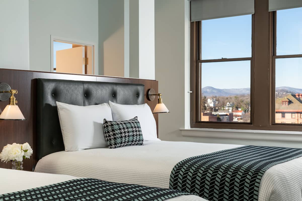 Una habitación de hotel con una cama grande, dos lámparas de noche, vistas a las montañas y una ventana con vista al pueblo.