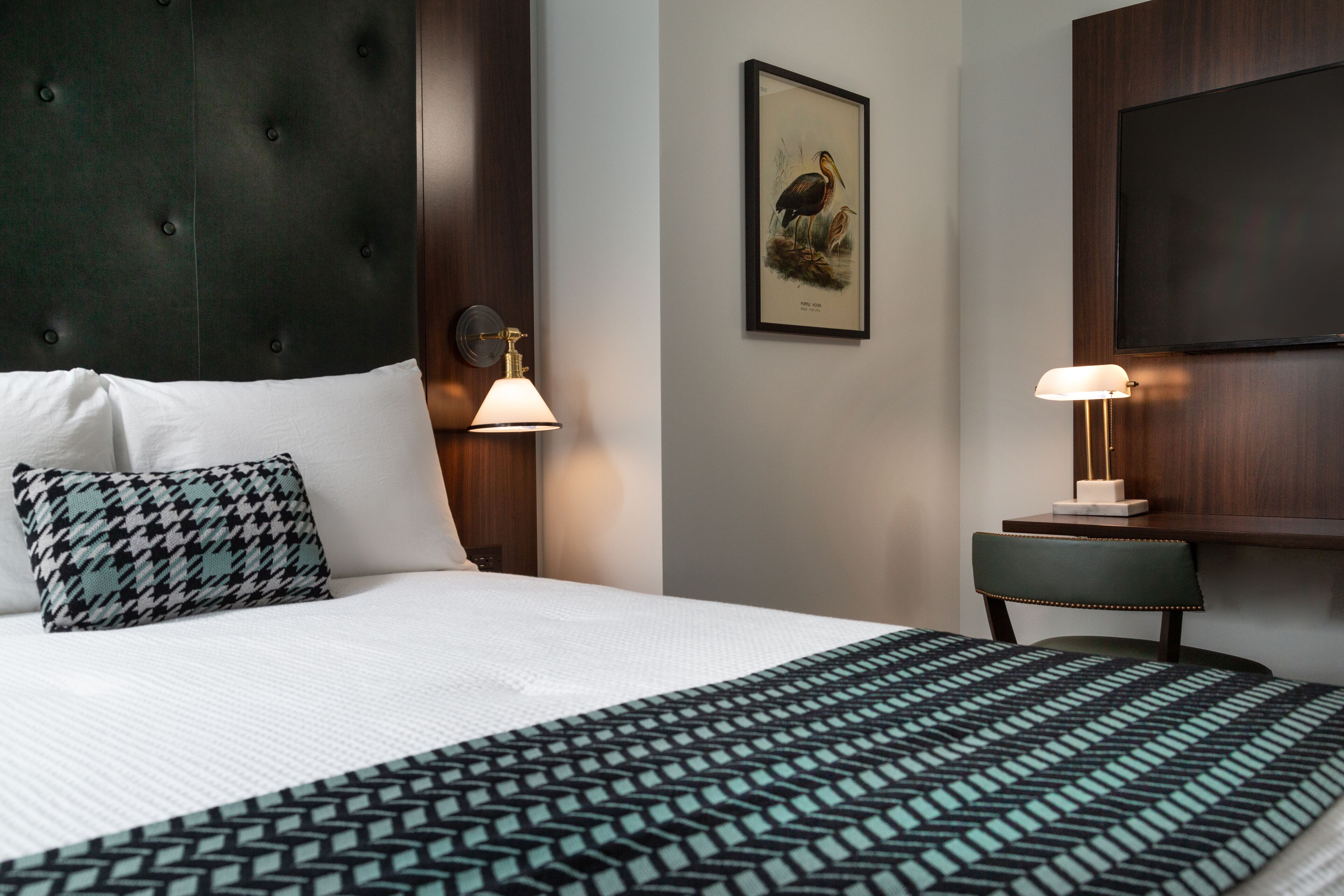 Un lit bien fait, recouvert d’une couvre-lit à motifs, agrémenté d’un coussin à carreaux et d’une lampe fixée au mur.