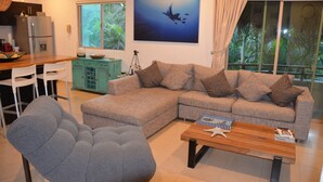 Una sala de estar moderna con un sofá seccional, un sillón gris y una mesa de centro de madera. Hay una zona de cocina con un refrigerador y una mesa de comedor con sillas. Un ventanal ofrece vistas a la vegetación exterior.