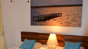 Habitación de hotel con dos camas, una mesita de noche con lámpara y un cuadro grande de un muelle sobre el agua.