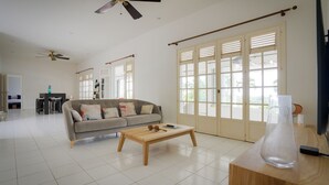 A spacious living area with a sofa, coffee table, and a TV on a wooden stand. There are ceiling fans, a dining area, and large windows with shutters.