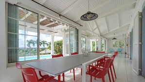 A spacious dining area with a long table and red chairs, large windows with a view of palm trees, and a ceiling with wooden beams.