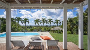 A covered outdoor area with a pool, lounge chairs, and a view of palm trees and greenery.