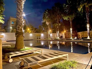 Zona de piscina nocturna con sillones de descanso y palmeras.