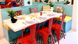 Un comedor moderno con una mesa blanca para cuatro personas, rodeada de sillas rojas. Hay un banco tapizado en azul con cojines, y un suelo estampado.