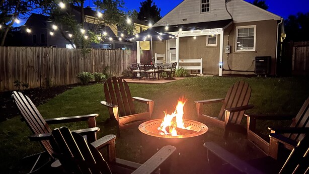 A backyard with a fire pit surrounded by Adirondack chairs, a wooden deck, and string lights. - The Cottage on Royal - Historic Leesburg Home (Leesburg)