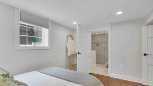 A bedroom with a bed, a window with blinds, a door leading to a bathroom, and a shower area. - The Cottage on Royal - Historic Leesburg Home (Leesburg)