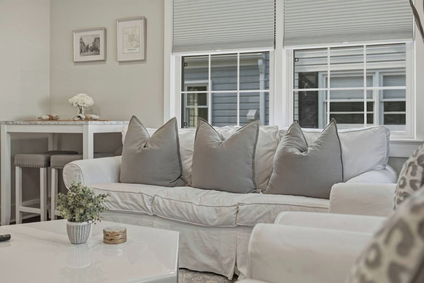 Ein Wohnzimmer mit einem weißen Sofa, einem Couchtisch und einem kleinen Beistelltisch mit Vase und Kerze. An der Wand hängen zwei gerahmte Bilder und ein Fenster mit Jalousien.