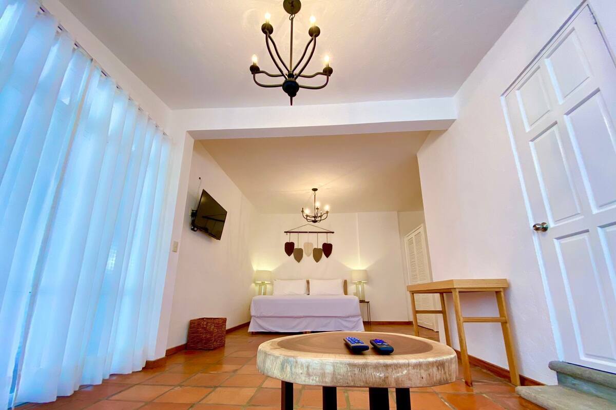 Una camera d'albergo con un letto, una TV fissata al muro, un lampadario e un piccolo tavolino rotondo con dei telecomandi.