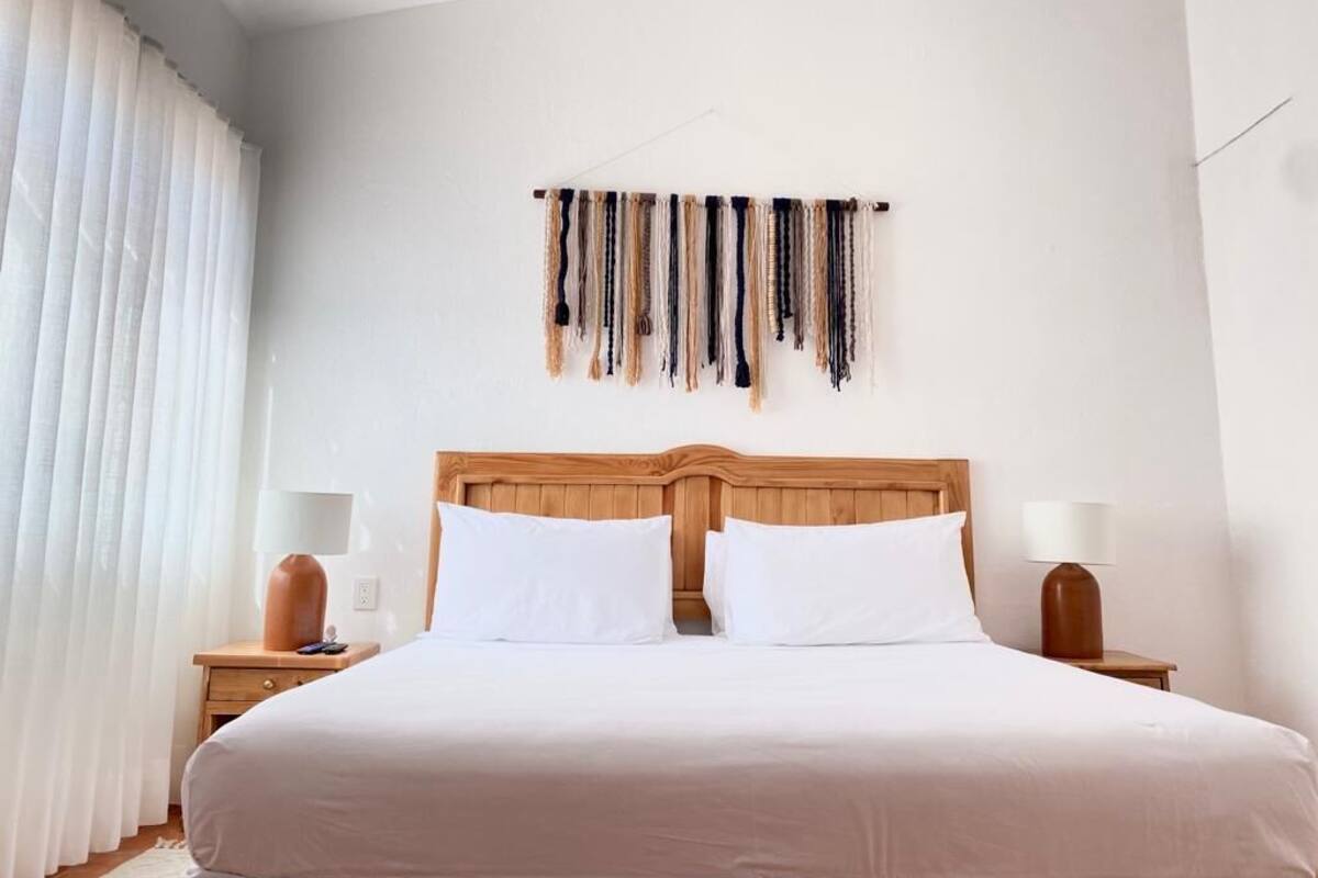 Una camera d'albergo con una testiera in legno, due cuscini bianchi, un comodino con una lampada e un arazzo appeso al muro.