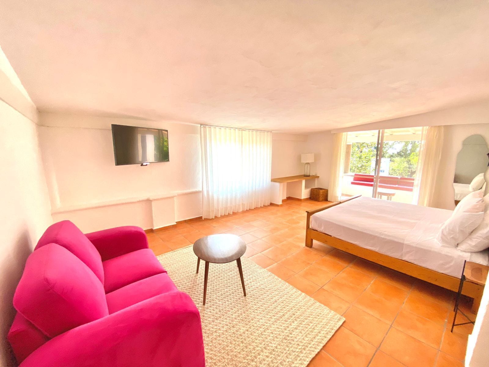 A bedroom with a bed, a TV mounted on the wall, a window with curtains, a small table with a lamp, and a bright orange tiled floor.