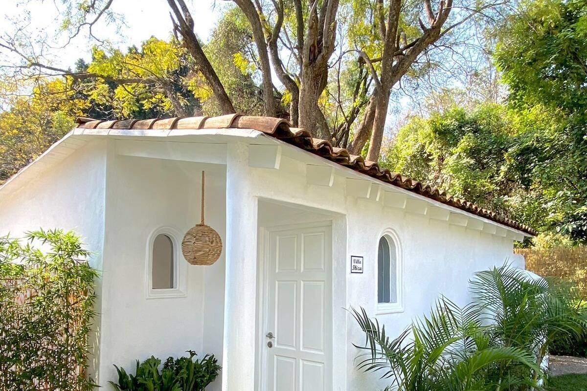 Un piccolo edificio a un piano con facciata bianca e tetto in tegole. Si nota una porta d'ingresso con un piccolo oblò sopra e un lampadario a sospensione. L'edificio è circondato da vegetazione, tra cui un recinto in bambù e diverse piante.