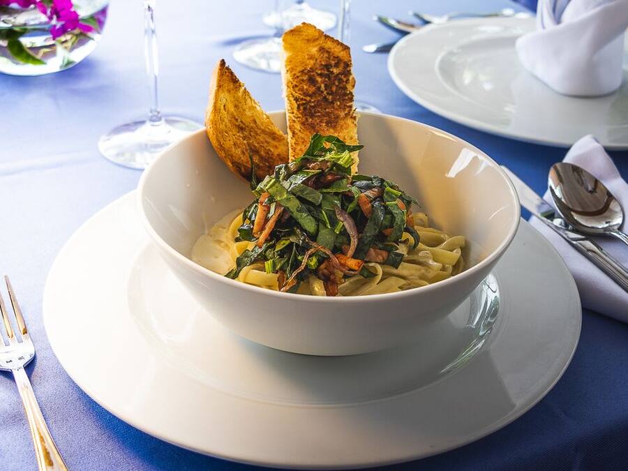Un plato de pasta con vegetales y tocino, servido en un plato blanco con un tenedor al lado.