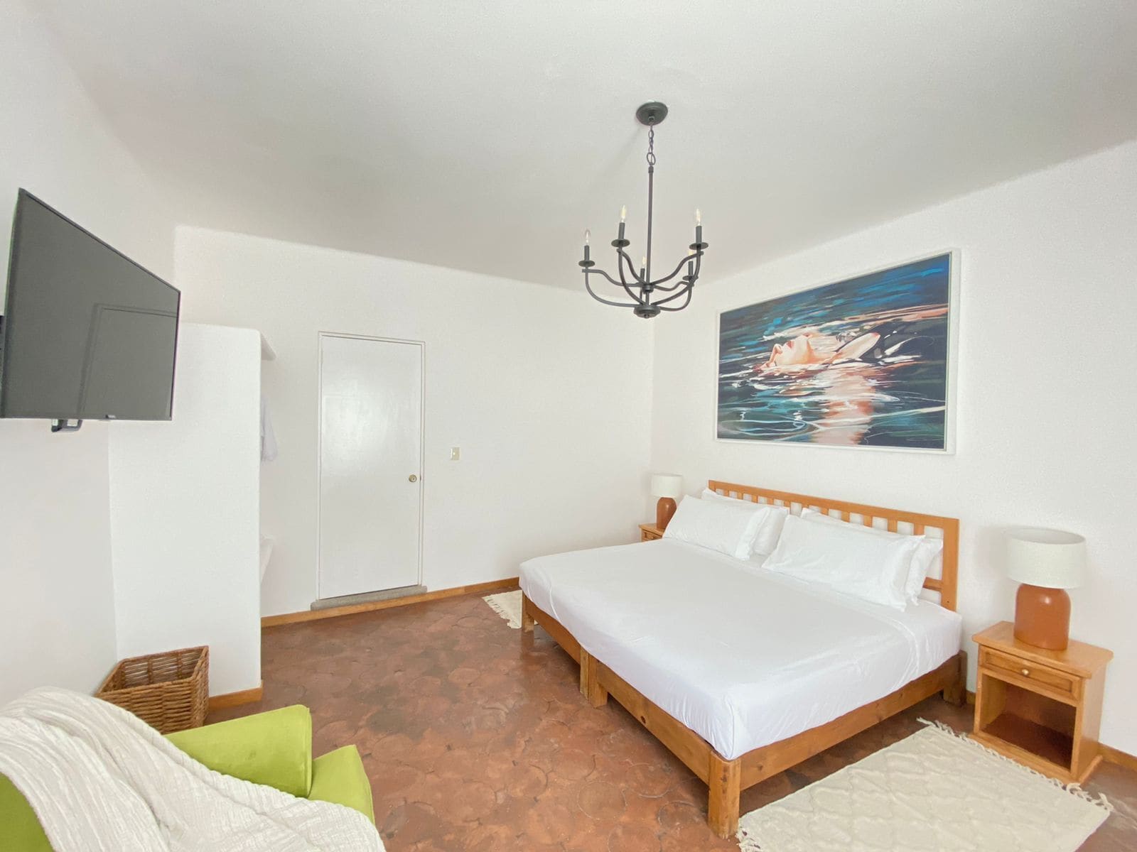 Een slaapkamer met een bed, een aan de muur bevestigde televisie, een schilderij en een nachtkastje van hout met een lamp.