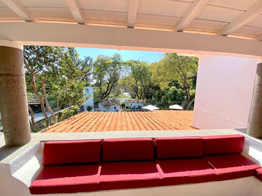A covered outdoor seating area with a red bench, a tiled roof, and a view of trees and buildings in the background.