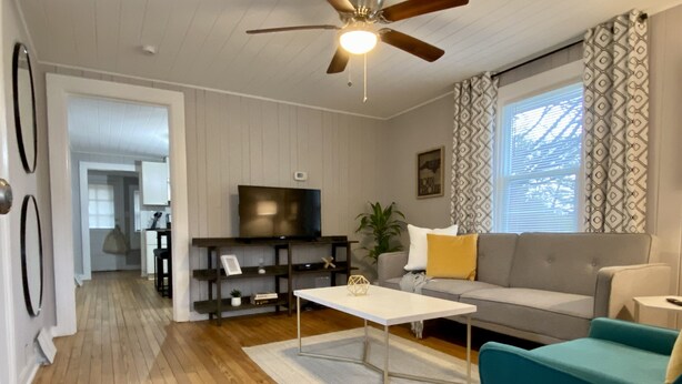 A living room with a wooden floor, a ceiling fan, a TV on a stand, a sofa, a coffee table, and a window with patterned curtains. - Quaint 2 bed 1 bath cottage style home (Kernersville)