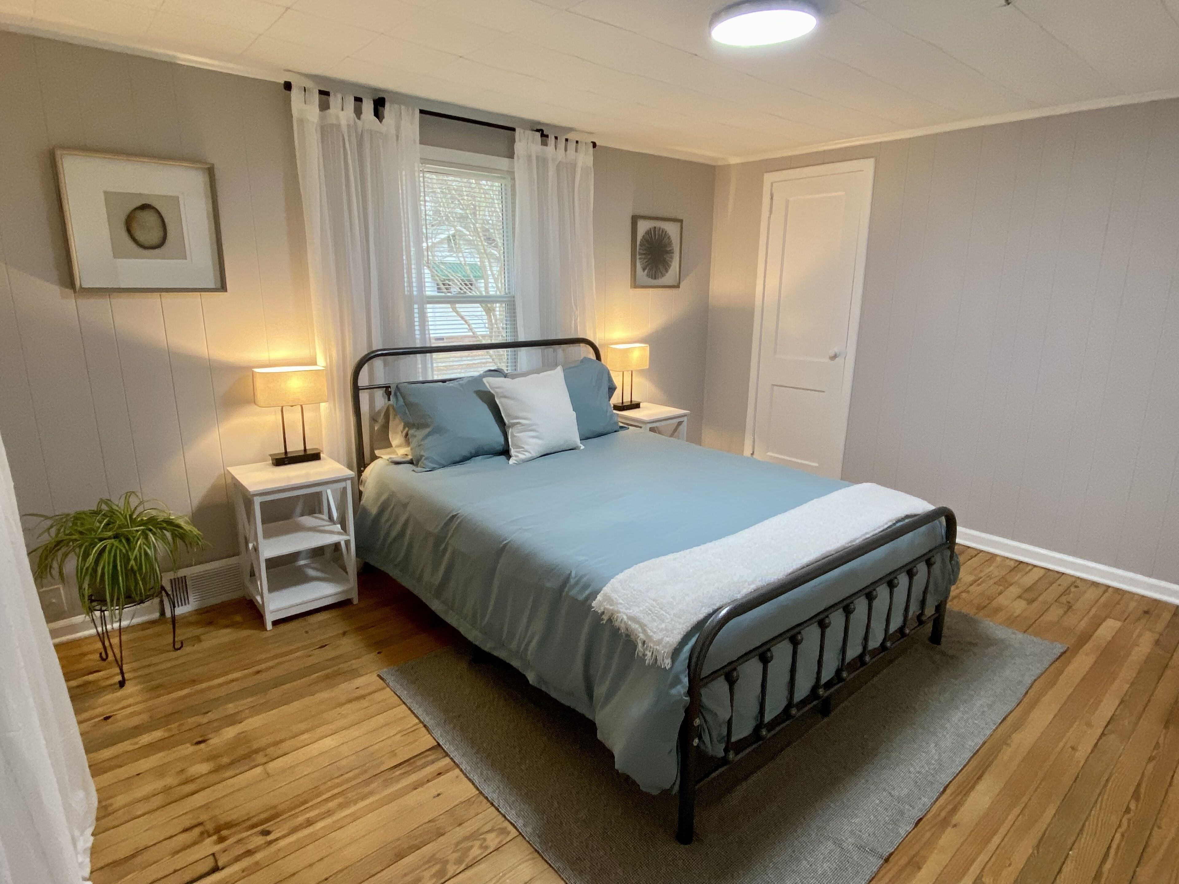 A neatly arranged bedroom with a metal bed frame, a bedside table with a lamp, a small plant, and a window with sheer curtains.