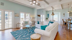 A spacious living room with a white sofa, a round coffee table, and a blue patterned rug. There is a ceiling fan, a dining area with a wooden table and chairs, and large windows allowing natural light.