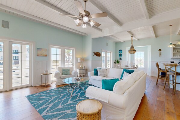 A spacious living room with a white sofa, a round coffee table, and a blue patterned rug. There is a ceiling fan, a dining area with a wooden table and chairs, and large windows allowing natural light.