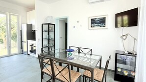 A modern kitchen with a built-in oven, a dining area with a glass table and wicker chairs, a TV mounted on the wall, and a small cabinet with dishes. - N122 - Numana, delightful single villa with garden (Numana)